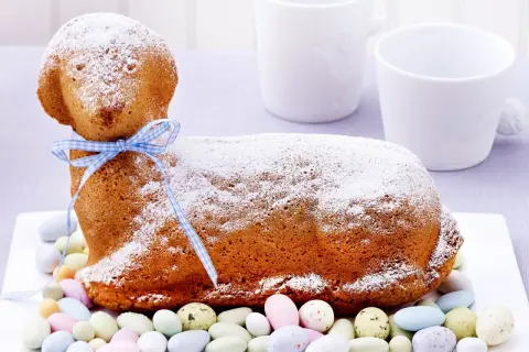Ein süßes Osterlamm-Rezept: Ein goldbrauner Kuchen in Lammform, mit Puderzucker bestäubt und einem blauen Band verziert, umgeben von bunten Zuckereiern.