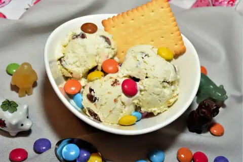 Schüssel mit cremigem Eis, garniert mit bunten Smarties und einem Keks. Kleine Figuren und weitere Smarties schmücken den Tisch.