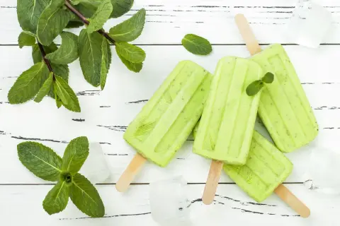 Erfrischendes Minzeis selber machen: Grüne Eis am Stiel mit frischer Minze und Eiswürfeln auf weißem Holzuntergrund.