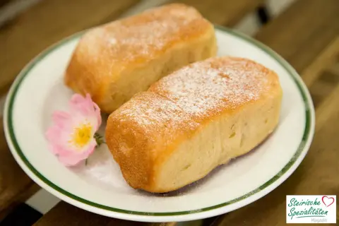 Zwei goldbraune Buchteln, mit Puderzucker bestäubt, auf einem Teller mit einer rosa Blüte. Ein köstliches Buchteln Rezept für Genießer.