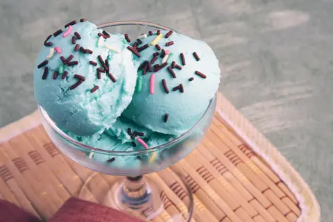 Ein Glas mit engelblauem Eis, bestreut mit Schokostreuseln und bunten Zuckerperlen, steht auf einem Bambusteller.