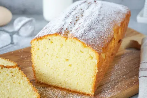 Ein einfacher Kuchen, bestäubt mit Puderzucker, liegt auf einem Holzbrett. Daneben liegen zwei Scheiben des Kuchens.