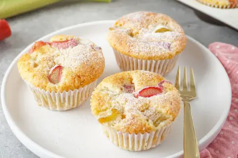 Drei saftige Rhabarber Muffins, mit Puderzucker bestäubt, auf einem weißen Teller. Ein goldener Gabel liegt daneben.