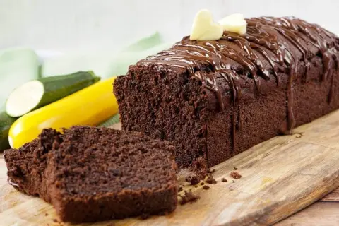 Saftiger Schokoladen-Zucchini-Kuchen mit Schokoladenglasur und zwei Herz-Dekorationen, daneben liegen frische Zucchini.