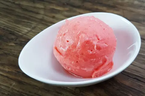 Ein Löffel cremiges Erdbeereis mit Sahne in einer weißen Schale auf Holzhintergrund.