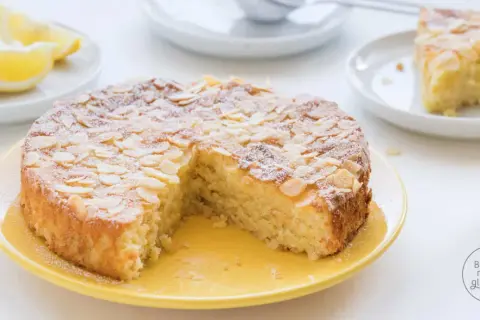 Saftiger mandelkuchen rezept, bestäubt mit Puderzucker und Mandelsplittern, daneben Zitronenspalten und eine ganze Zitrone.