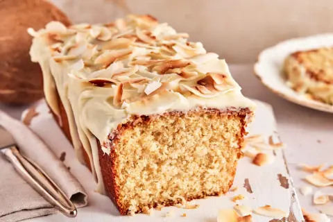 Ein saftiger Kokos Kuchen mit weißer Glasur und gerösteten Kokoschips. Daneben liegt eine Kokosnuss.