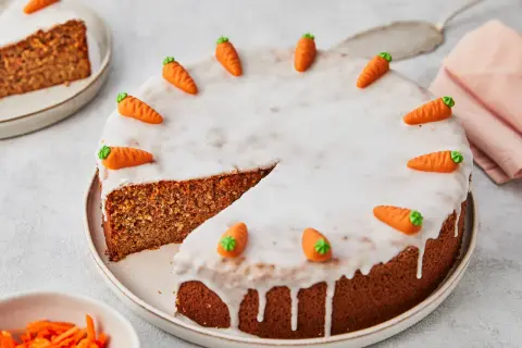 Ein saftiger karottenkuchen ohne mehl, verziert mit Zuckermöhren und Zuckerguss. Ein Stück ist bereits angeschnitten.
