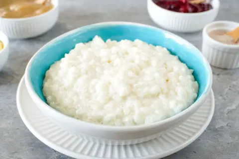Cremiger Milchreis im blauen Schälchen, daneben Schüsseln mit Apfelmus, Kirschen und Zimt. Perfekt für ein einfaches Milchreis Rezept.