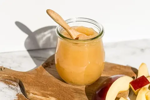 Glas mit selbstgemachtem Apfelmus, Holzlöffel und Apfelstücke auf einem Holzbrett mit Zimt.