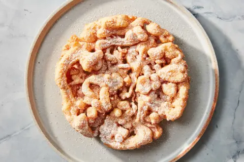 Ein goldbrauner, puderzuckerbestäubter **Funnel Cake** auf einem grauen Teller.