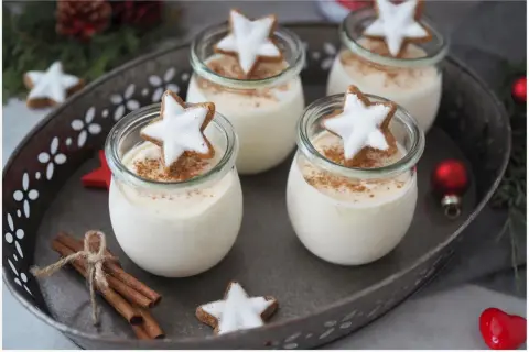 Vier Gläser mit cremigen Parfait-Rezepten im Glas, garniert mit Zimt und Sternenplätzchen. Weihnachtliche Deko mit Zimtstangen und Kugeln.