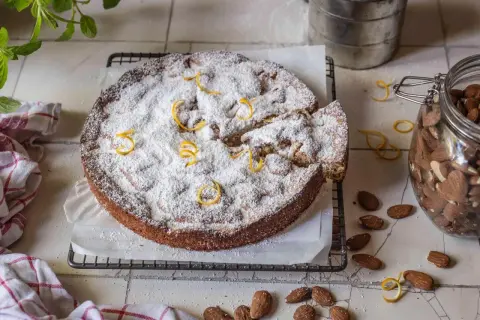 Ein mallorquinischer Mandelkuchen, bestäubt mit Puderzucker und garniert mit Zitronenzesten, neben einem Glas Mandeln.