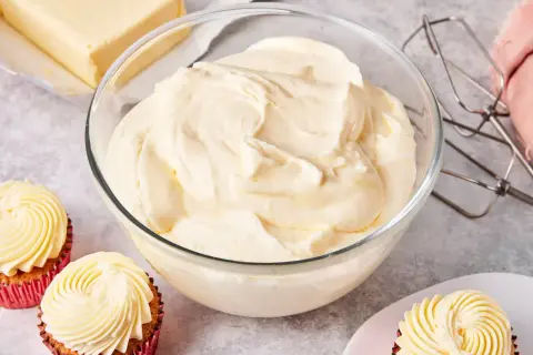 Schüssel mit cremiger amerikanischer Buttercreme, daneben ein Block Butter, Cupcakes und ein Handmixer.