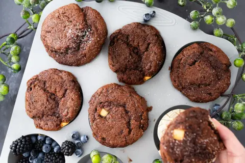 Fünf schoko muffins mit weißen Schokostückchen und Puderzucker in einer Muffinform, daneben Beeren und grüne Beeren.