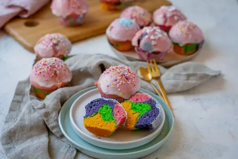 Bunte regenbogen muffins mit rosa Zuckerguss und Streuseln, ein Stück auf einem Teller zeigt das farbenfrohe Innere.