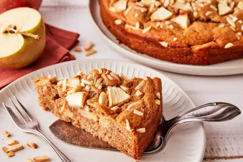Ein Stück saftiger apfelkuchen ohne zucker, garniert mit Mandelblättchen. Daneben ein halber Apfel und eine Gabel.