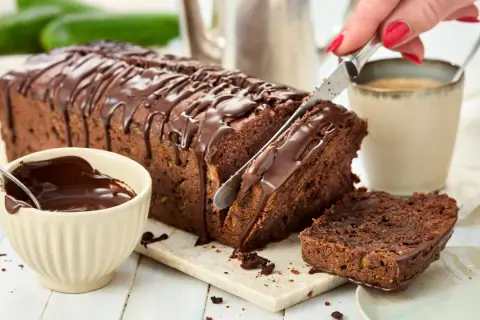 Ein saftiger Zucchini Schoko Kuchen wird angeschnitten. Daneben eine Schale mit Schokoladenglasur und eine Tasse Kaffee.