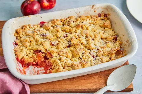 Ein frisch gebackener pflaumen crumble in einer weißen Auflaufform, bereit zum Servieren. Daneben liegen zwei reife Pflaumen.