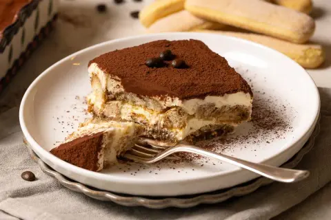 Ein Stück Tiramisu, das nach einem einfachen tiramisu rezept gemacht wurde, liegt auf einem Teller. Kakaopulver bestäubt die cremige Schicht und die Löffelbiskuits.