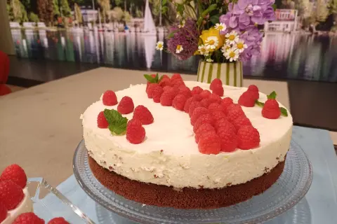 Himbeer-Mascarpone-Torte mit frischen Beeren und Minze, dekoriert mit einem Blumenstrauß im Hintergrund.