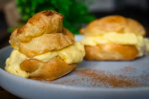 Zwei köstliche Windbeutel, gefüllt mit cremiger Vanillecreme, auf einem Teller mit Zimt bestäubt. Ein perfektes Windbeutel Rezept zum Nachmachen.