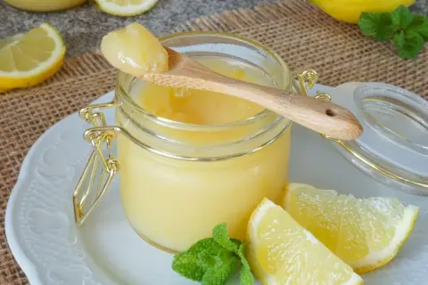 Ein Glas mit cremigem Lemon Curd, garniert mit Zitronenspalten und Minze. Ein Holzlöffel mit einer Portion Lemon Curd ruht auf dem Glas.