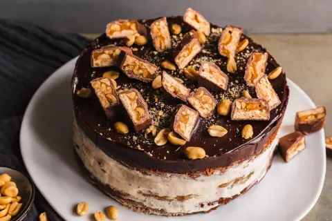Eine köstliche Snickers Torte mit Schokoladenglasur, Erdnüssen und vielen Snickers-Stücken obendrauf.