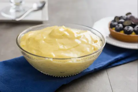 Cremige Crema Pasticcera in einer Glasschüssel, verfeinert mit Vanille. Daneben ein Törtchen mit Blaubeeren.