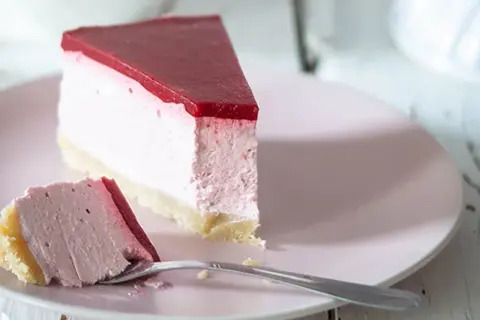 Ein Stück cremiger cheesecake ohne backen mit roter Fruchtschicht auf rosa Teller, daneben ein Gabel mit einem Stück Kuchen.