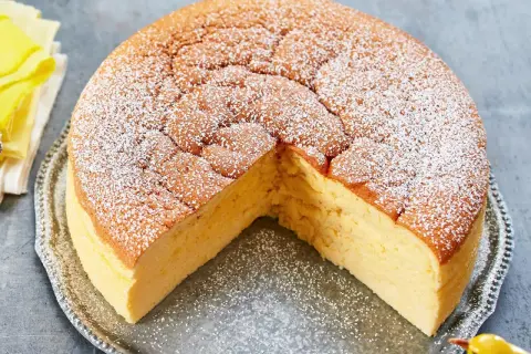Ein fluffiger, goldbrauner low carb kuchen, bestäubt mit Puderzucker, auf einer silbernen Platte. Ein kleines gelbes Vogel-Dekoration sitzt daneben.