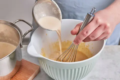 Eine cremige Masse wird aus einem Topf in eine Schüssel gegossen, während mit einem Schneebesen gerührt wird. Perfekt für selbstgemachtes Zimteis aus der Eismaschine.