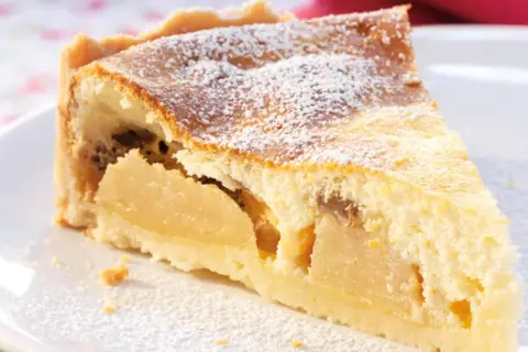 Ein Stück Apfelkuchen mit Quark, bestäubt mit Puderzucker, auf einem weißen Teller.
