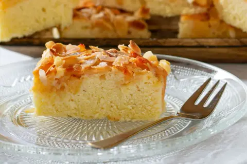 Ein saftiges Stück mandelkuchen vom blech, bestreut mit gerösteten Mandeln und Zucker, liegt auf einem Glas-Teller.