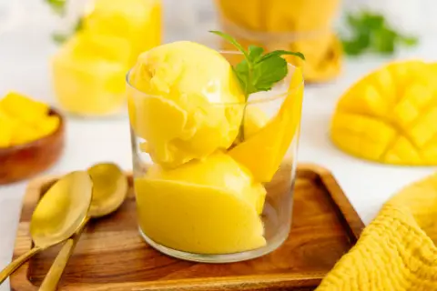 Erfrischendes Mango Eis in einem Glas, garniert mit Minze und Mangostücken. Daneben liegen goldene Löffel auf einem Holztablett.