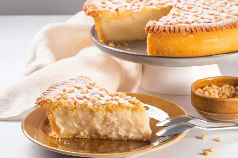 Ein Stück Torta della Nonna, bestäubt mit Puderzucker und Pinienkernen, auf einem Teller neben einer Schüssel voller Pinienkerne.