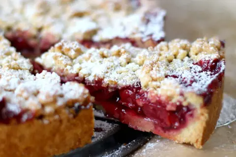Saftiger kirschkuchen mit streusel, bestäubt mit Puderzucker. Ein Stück wurde herausgeschnitten, um die roten Kirschen zu enthüllen.