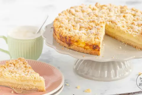 Ein saftiger Streuselkuchen mit Puddingfüllung, bestäubt mit Puderzucker. Daneben eine Tasse Kaffee und ein Stück Kuchen auf einem Teller.