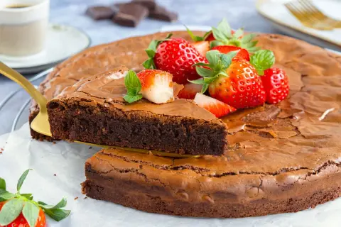 Ein saftiger brownie kuchen, garniert mit frischen Erdbeeren und Minze. Ein Stück wird mit einem goldenen Tortenheber herausgehoben.