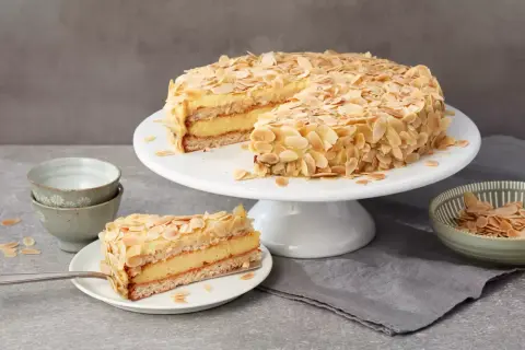Eine köstliche schwedische Mandeltorte, üppig mit Mandelblättchen bedeckt, wird auf einem Tortenständer präsentiert. Ein Stück ist bereits auf einem Teller angerichtet.