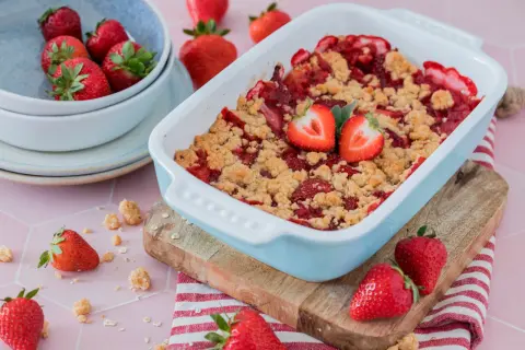 Ein frisch gebackener Erdbeer Crumble in einer hellblauen Auflaufform, garniert mit Erdbeerhälften. Frische Erdbeeren liegen daneben.