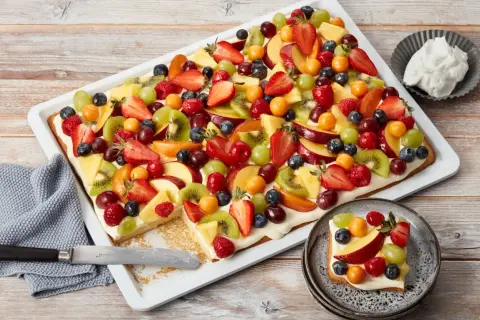 Bunter Blechkuchen mit Obst, belegt mit Erdbeeren, Kiwi, Trauben, Pfirsichen und Beeren. Ein Stück ist bereits auf einem Teller angerichtet.