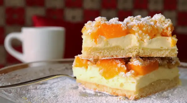 Zwei Stücke Mandarinen Schmand Kuchen Blech auf einem Teller, bestäubt mit Puderzucker. Im Hintergrund eine weiße Tasse.