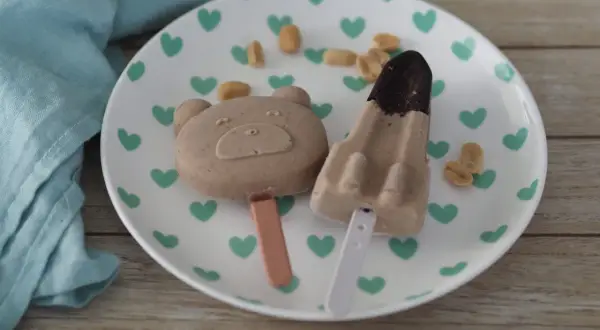 Zwei niedliche Eis am Stiel, ein Bär und eine Rakete, mit Erdnussbutter-Geschmack, liegen auf einem Teller mit grünen Herzen.