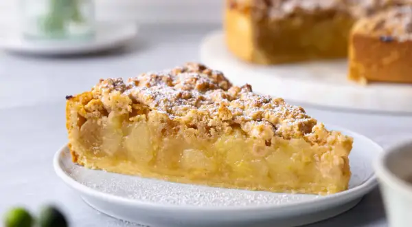 Ein saftiges Stück veganer Apfelkuchen mit Streuseln und Puderzucker bestäubt. Der Kuchen liegt auf einem weißen Teller, im Hintergrund ist ein ganzer Kuchen zu sehen.