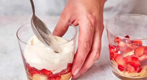 Eine Hand schichtet mit einem Löffel cremiges Topping auf ein Erdbeer-Tiramisu im Glas.