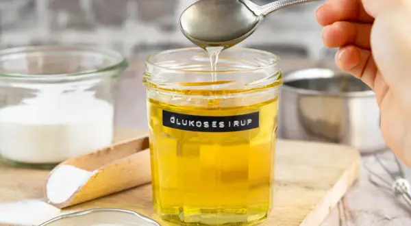 Ein Glas mit selbstgemachtem Glukosesirup, der von einem Löffel tropft. Zutaten wie Zucker sind daneben zu sehen.