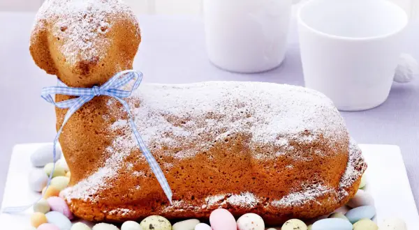 Ein süßes Osterlamm-Rezept: Ein goldbrauner Kuchen in Lammform, mit Puderzucker bestäubt und einem blauen Band verziert, umgeben von bunten Zuckereiern.