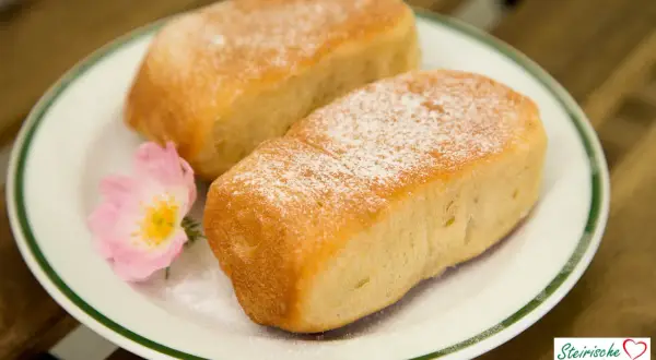 Zwei goldbraune Buchteln, mit Puderzucker bestäubt, auf einem Teller mit einer rosa Blüte. Ein köstliches Buchteln Rezept für Genießer.