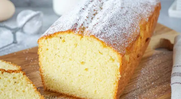 Ein einfacher Kuchen, bestäubt mit Puderzucker, liegt auf einem Holzbrett. Daneben liegen zwei Scheiben des Kuchens.